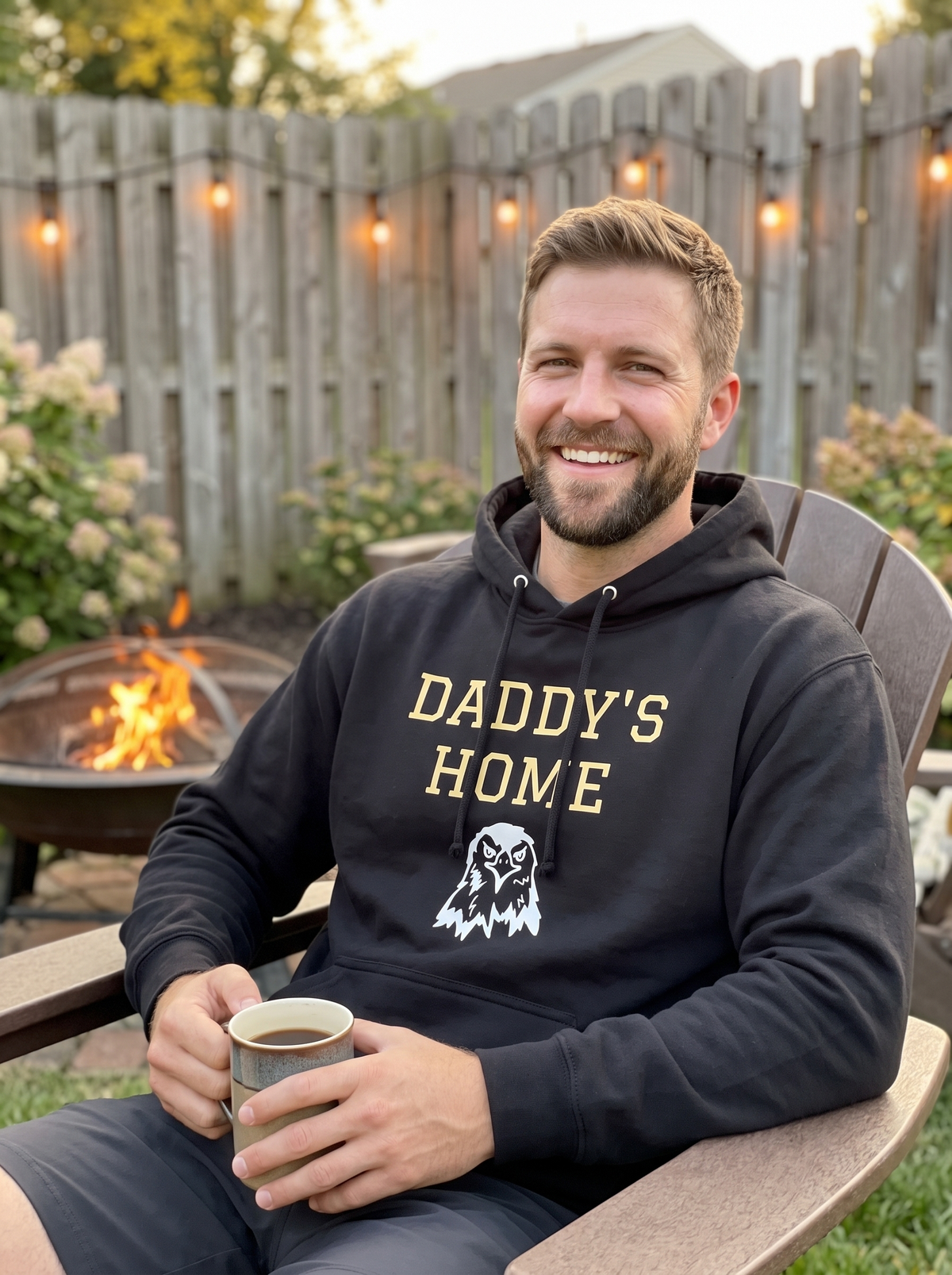 Man wearing the black Daddy's Home Vintage Heritage Hoodie with gold varsity lettering and eagle graphic, sitting by a backyard fire pit