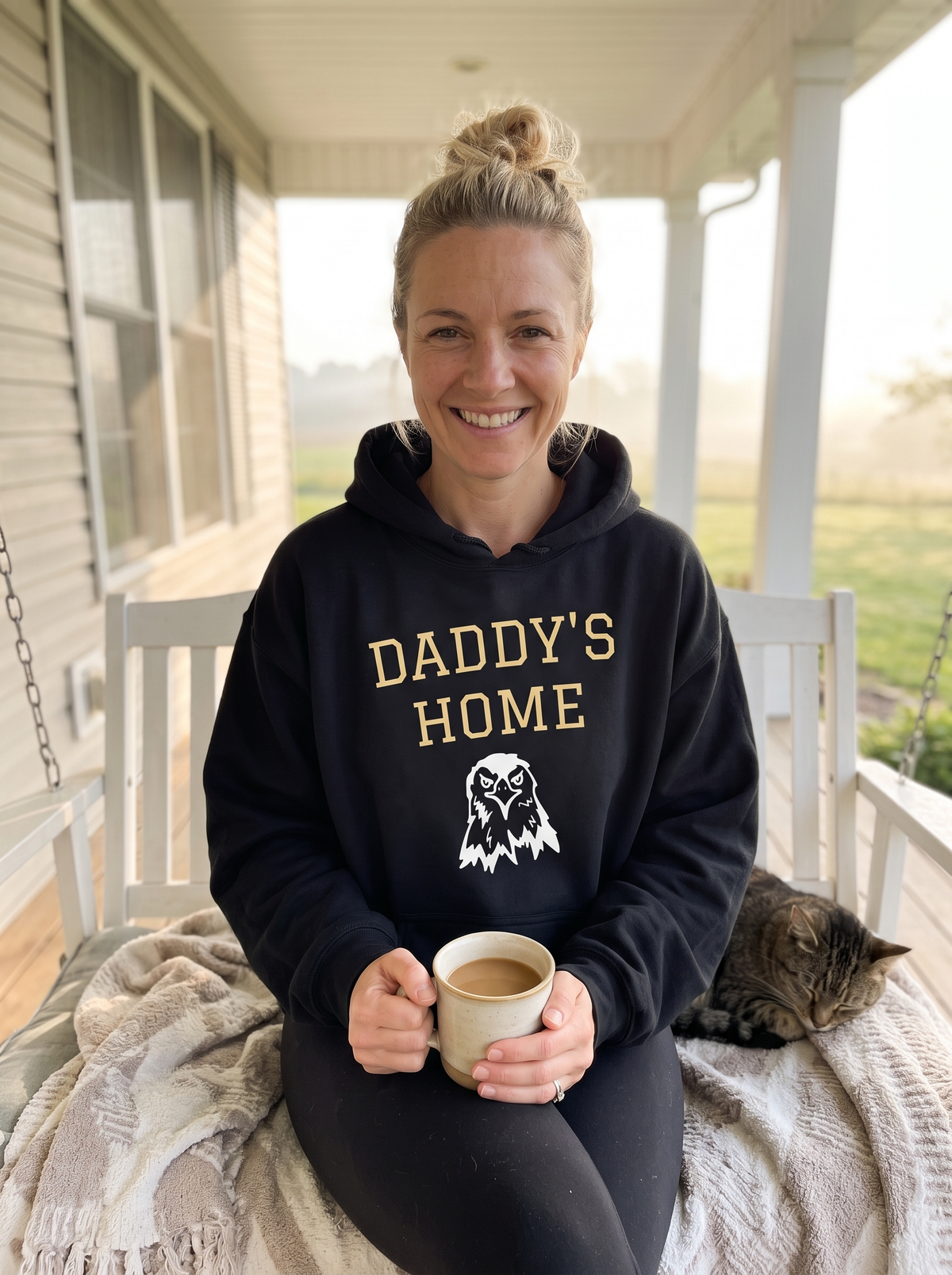 Woman wearing the oversized black Daddy's Home Vintage Heritage Hoodie with gold varsity lettering and eagle graphic on a farmhouse porch swing
