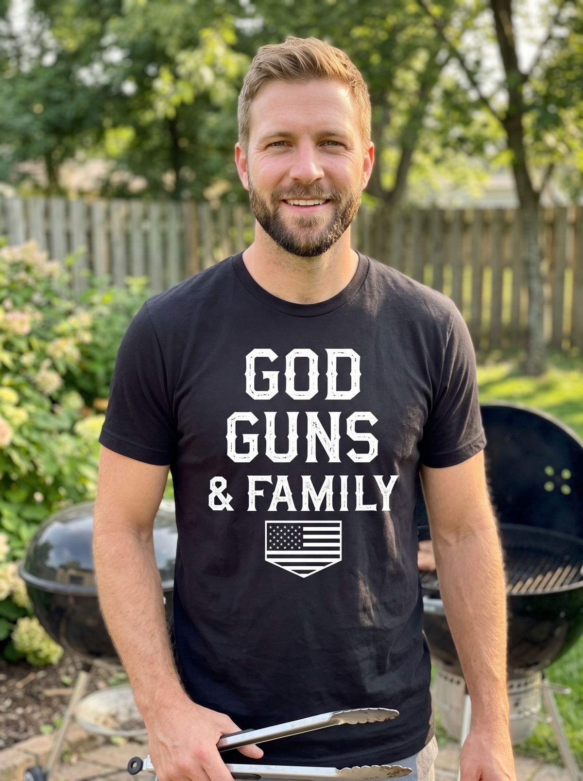 Man wearing the black heather God, Guns, & Family Classic Tee with white lettering and American flag shield, standing at a backyard grill