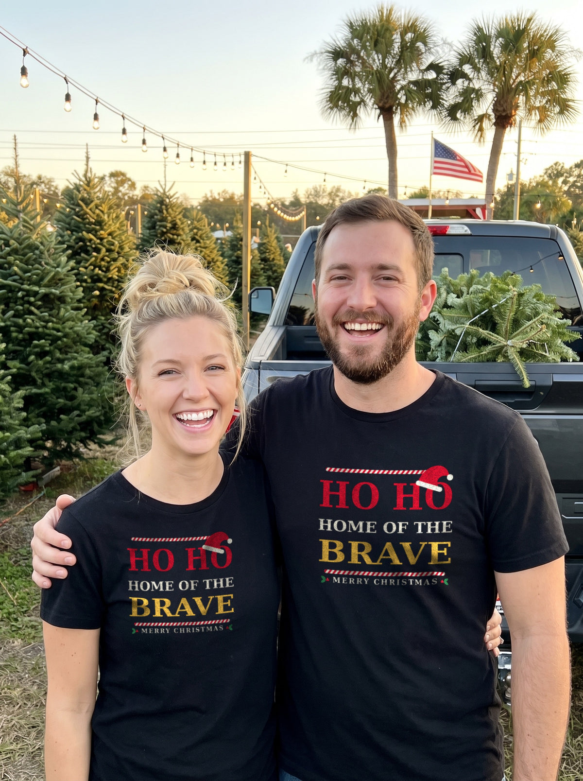 Garrett and Savannah in matching black "Ho Ho Home of the Brave" Christmas Patriot tees at a Christmas tree lot with an American flag
