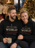 AI-generated photo of a couple in matching black Joy to the World Faith Family Freedom hoodies by a decorated Christmas tree and fireplace