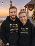 Couple in black Keep Christ in Christmas hoodies on a boardwalk at dusk