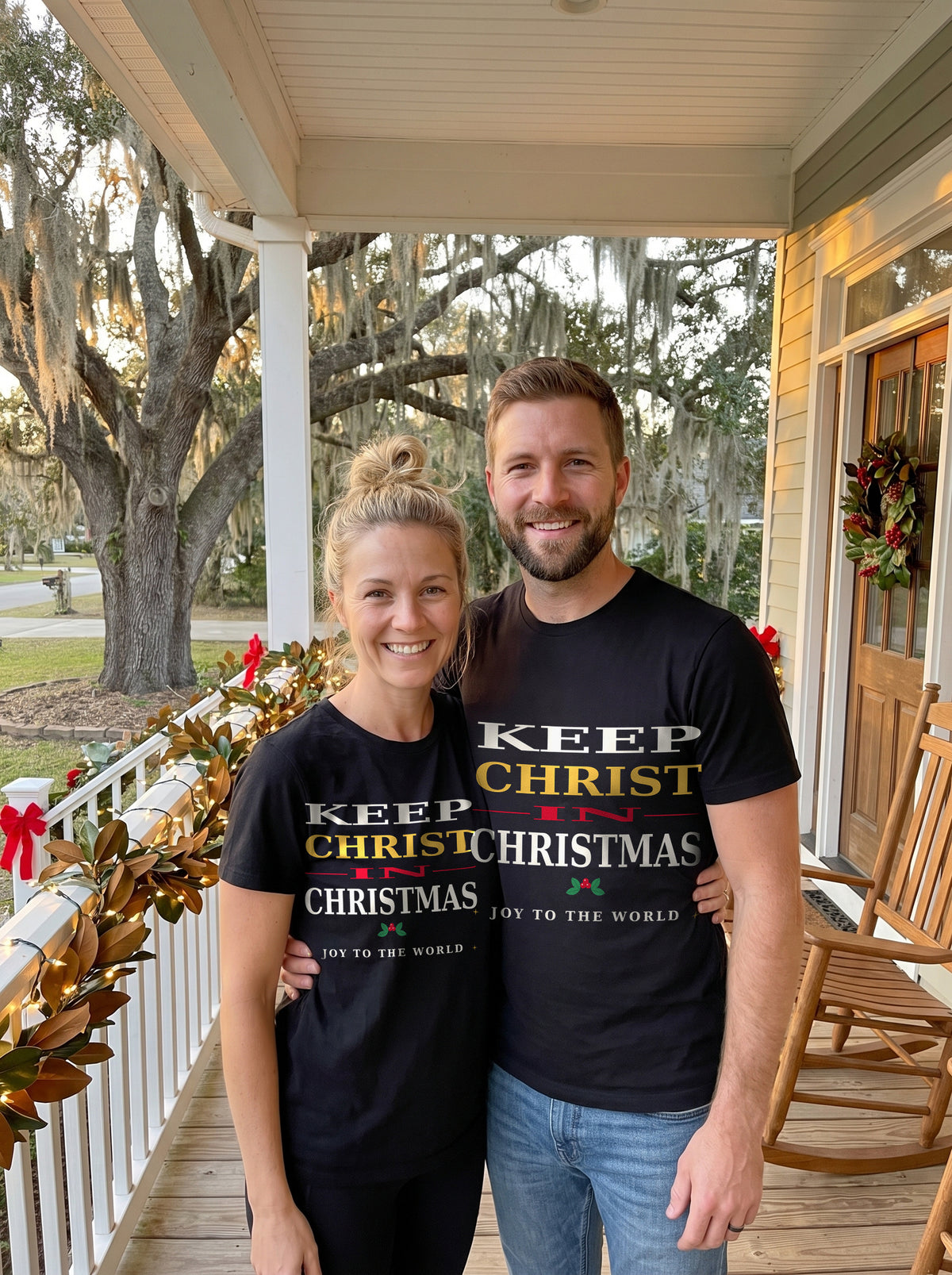 Couple in black Keep Christ in Christmas tees on a Southern front porch with magnolia garland and string lights at golden hour