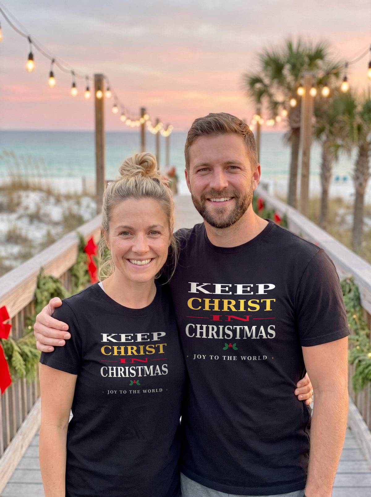 Couple in black Keep Christ in Christmas tees on a Gulf Coast beach boardwalk at dusk with string lights and holiday garland