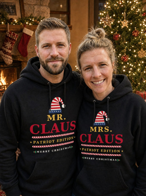 Garrett and Savannah wearing matching Mr. Claus and Mrs. Claus Patriot Edition black Christmas hoodies in front of a lit Christmas tree and fireplace
