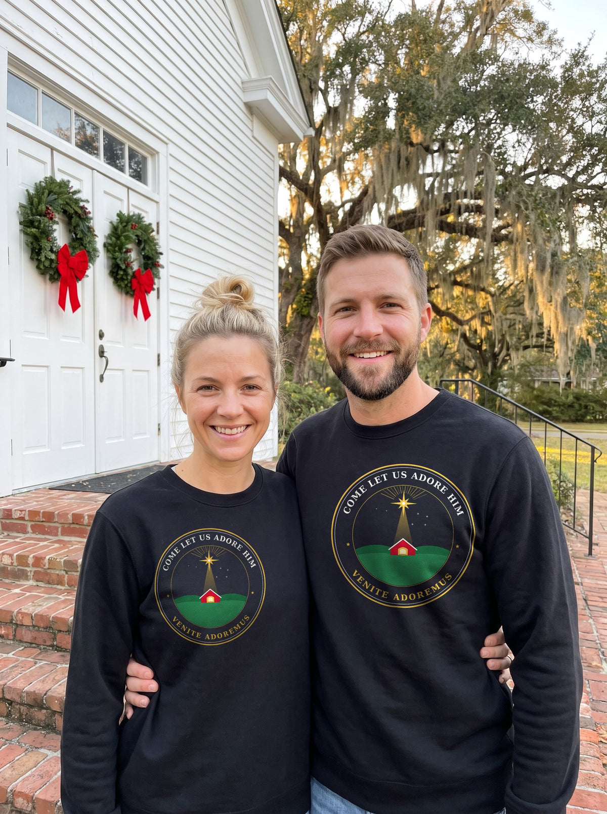 Man and woman in black Come Let Us Adore Him Nativity badge sweatshirts standing on church steps with Christmas wreaths
