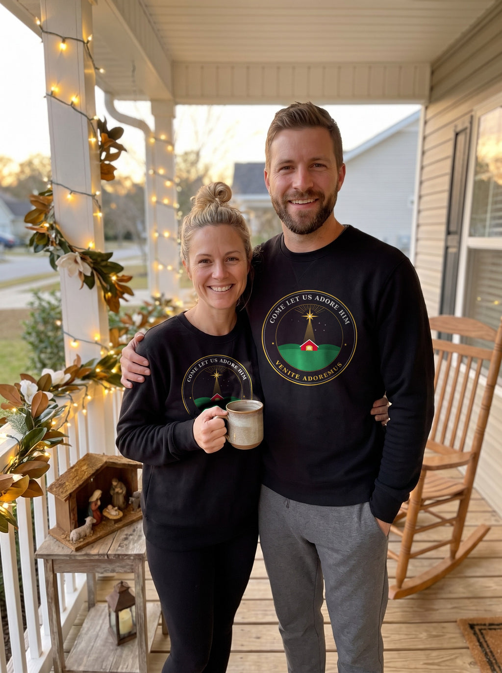 Couple in matching black Come Let Us Adore Him Nativity badge sweatshirts on a porch decorated with Christmas lights and a nativity scene