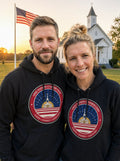 Garrett and Savannah wearing matching black One Nation Under God badge hoodies in front of a white country church with an American flag at sunrise
