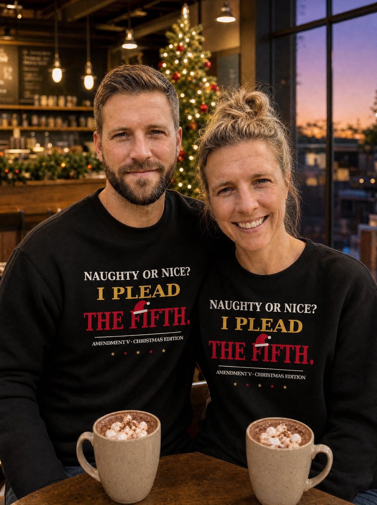Garrett and Savannah in matching black I Plead the Fifth Christmas Edition sweatshirts with a Santa hat graphic, holding hot cocoa in a decorated coffee shop at dusk