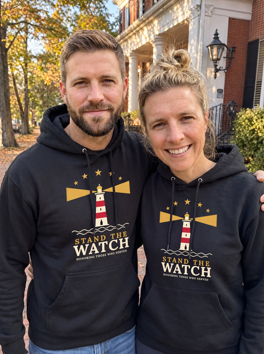 Couple wearing matching black Stand the Watch lighthouse hoodies on an autumn street (AI-generated image)
