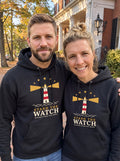Couple wearing matching black Stand the Watch lighthouse hoodies on an autumn street (AI-generated image)