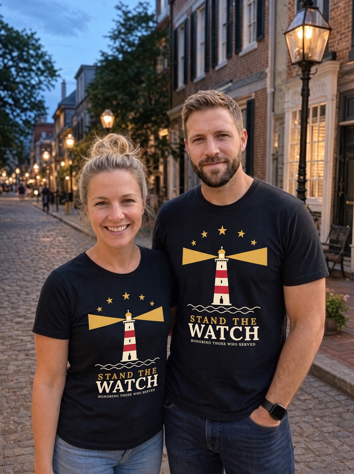 Couple wearing matching black Stand the Watch lighthouse tees on a cobblestone street at dusk (AI-generated image)